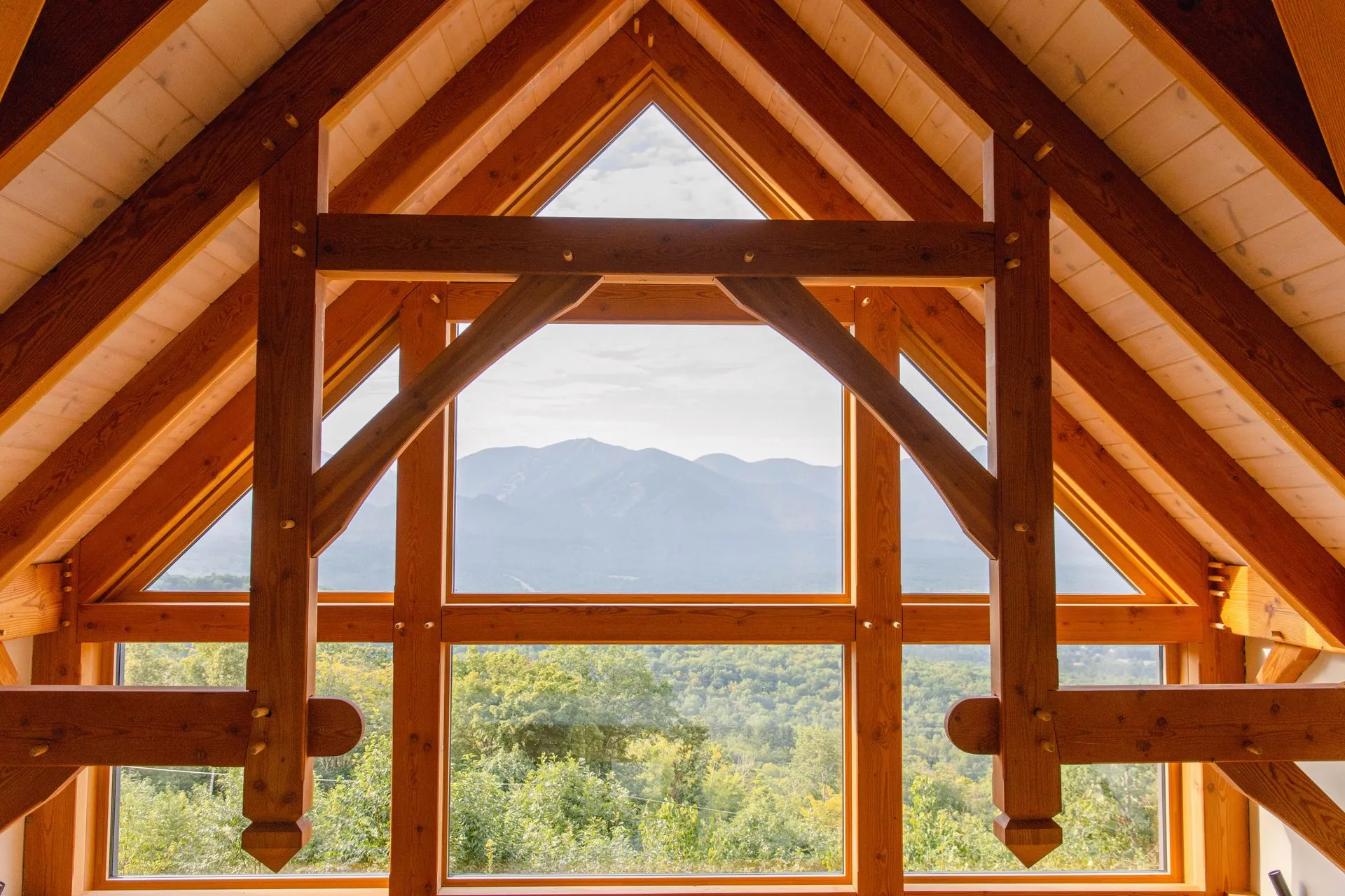 L'intérieur d'un comble : une charpente de bois clair, ferme triangulaire et pannes apparentes, encadre une grande baie vitrée ouverte sur une chaîne de montagnes boisées.