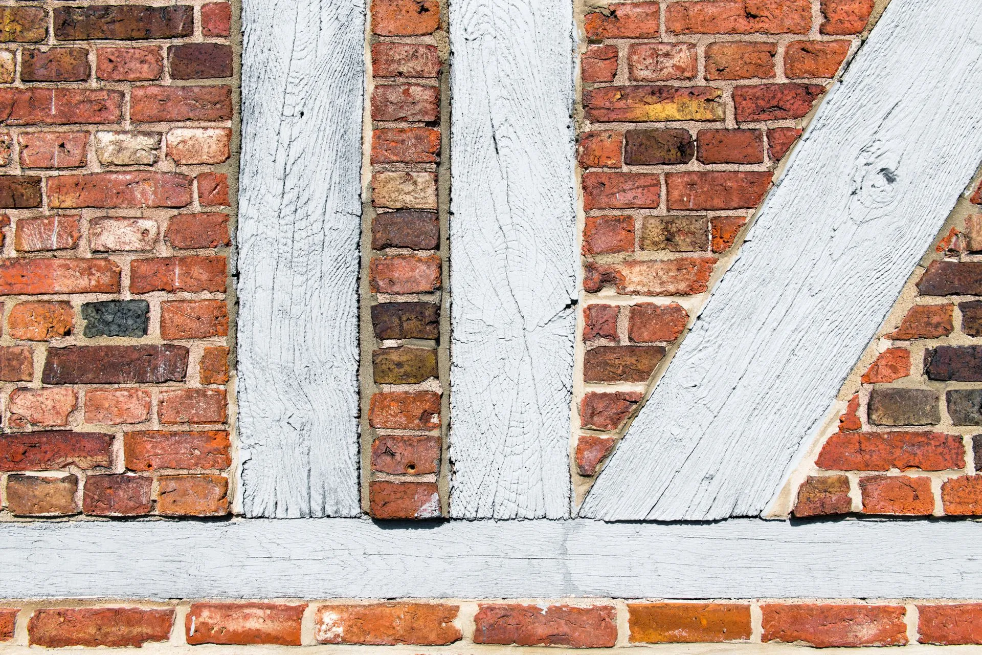 Un mur à colombage vu de face : des poutres de bois peintes en blanc, verticales et obliques, et entre elles un remplissage de briques rouges de teintes mêlées.