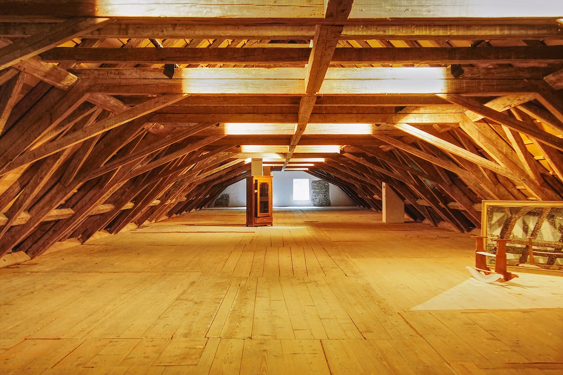 Le grenier vidé d'une vieille bâtisse : une charpente de bois massive à contrefiches, un plancher de larges lattes, une armoire vitrée isolée au fond près d'une petite fenêtre, un grand cadre et une chaise à bascule posés sous la pente à droite.