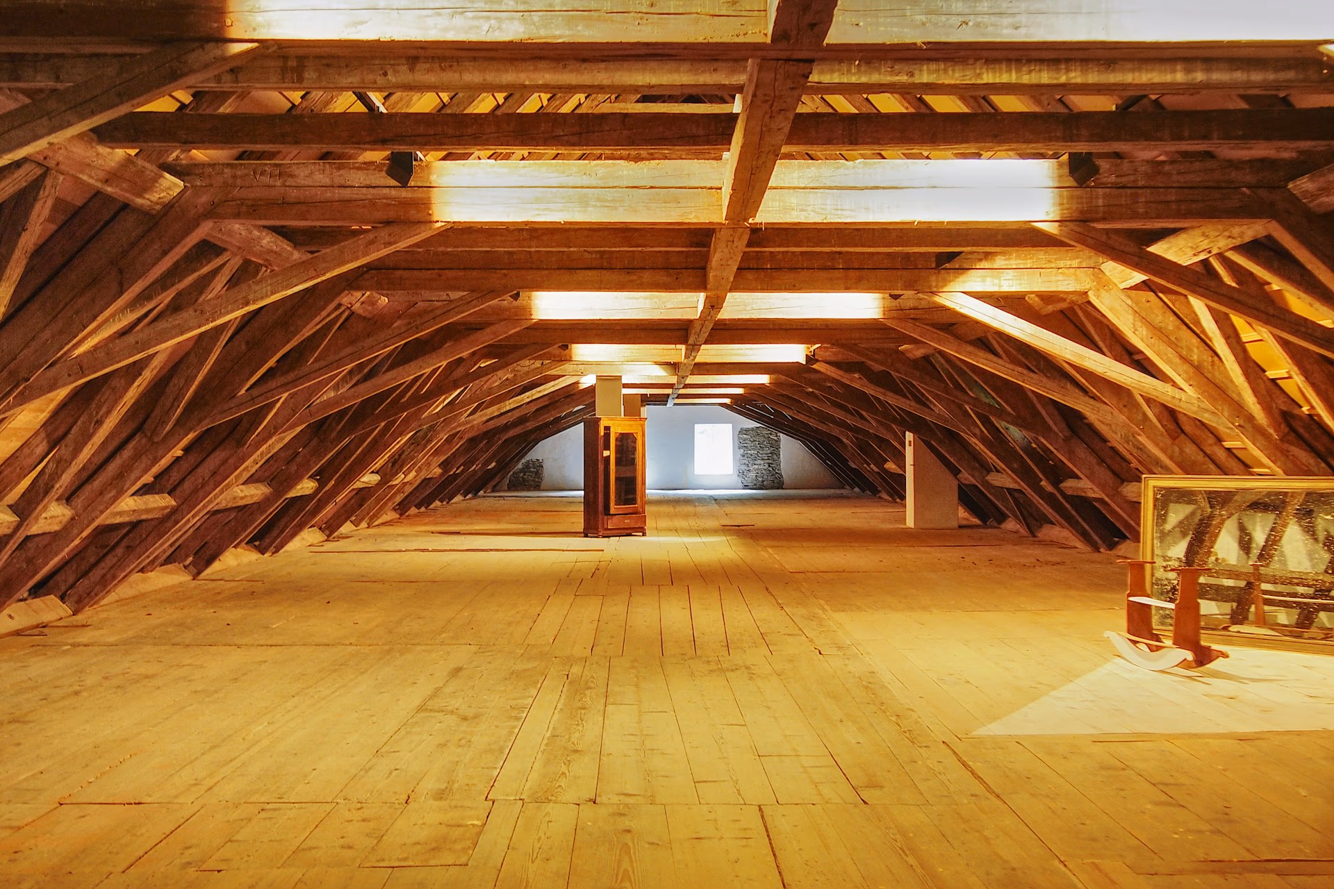 Le grenier vidé d'une vieille bâtisse : une charpente de bois massive à contrefiches, un plancher de larges lattes, une armoire vitrée isolée au fond près d'une petite fenêtre, un grand cadre et une chaise à bascule posés sous la pente à droite.