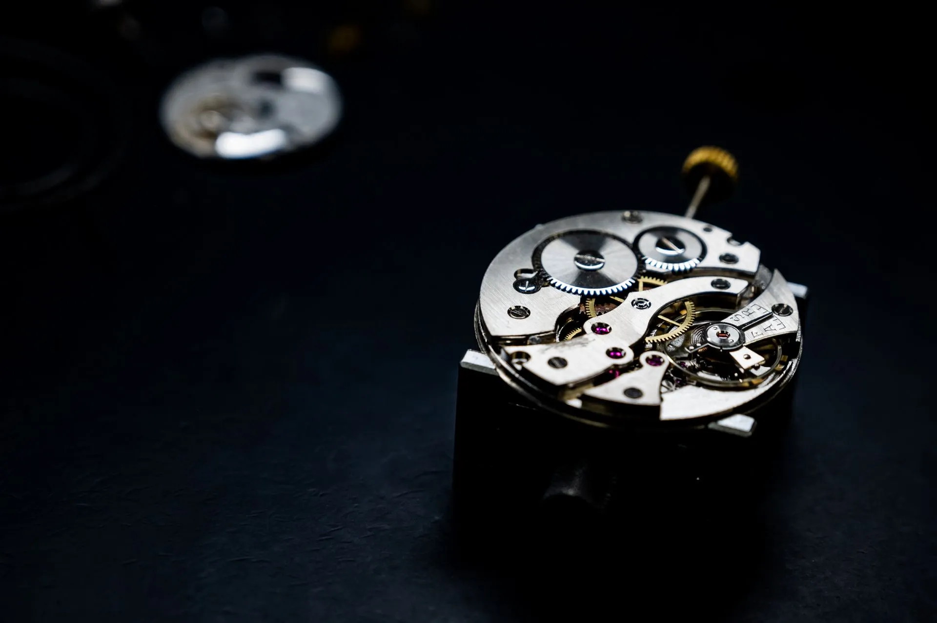 Un mouvement de montre mécanique posé sur un socle noir, vu de très près : des ponts d'acier brossé, des roues dentées dorées, quelques rubis rouges sertis dans les ponts et la tige de remontoir dressée sur le côté.