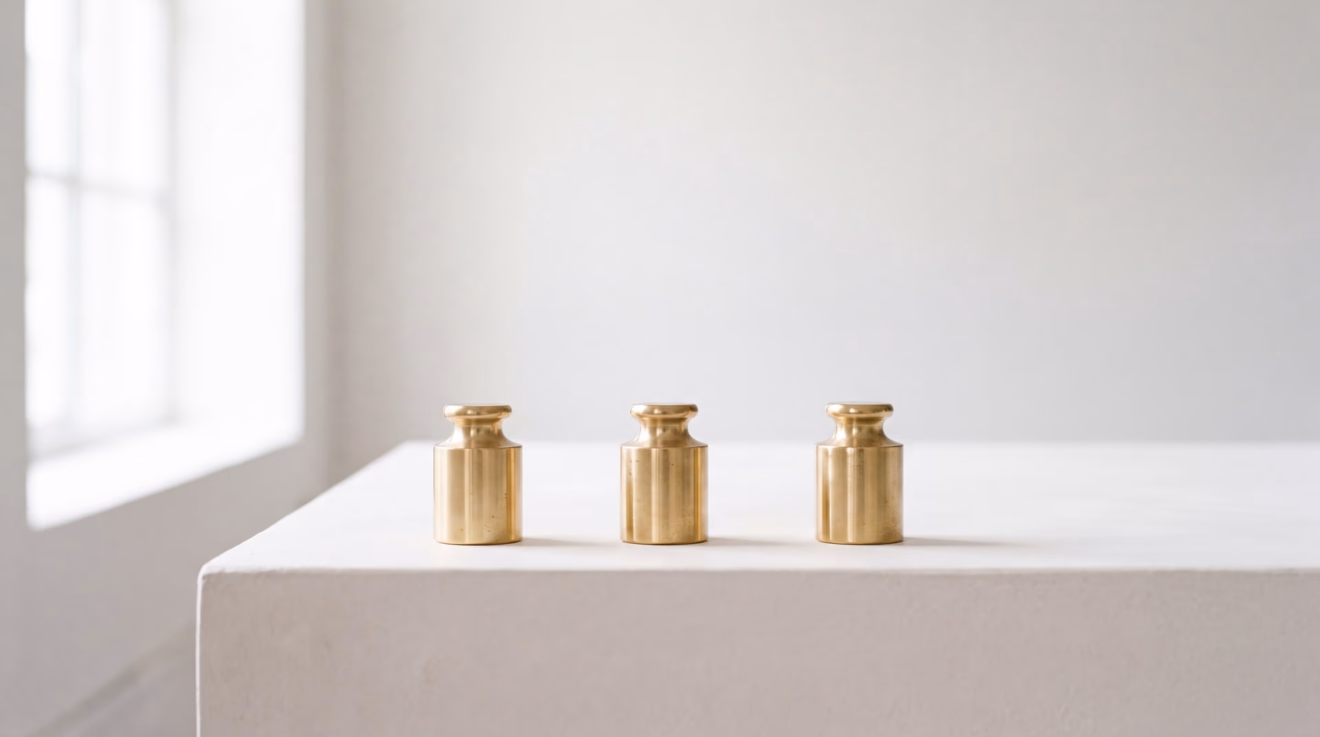 Trois poids étalons en laiton alignés sur un plan de plâtre clair, éclairés par la lumière d'une fenêtre, chacun posant une ombre courte.