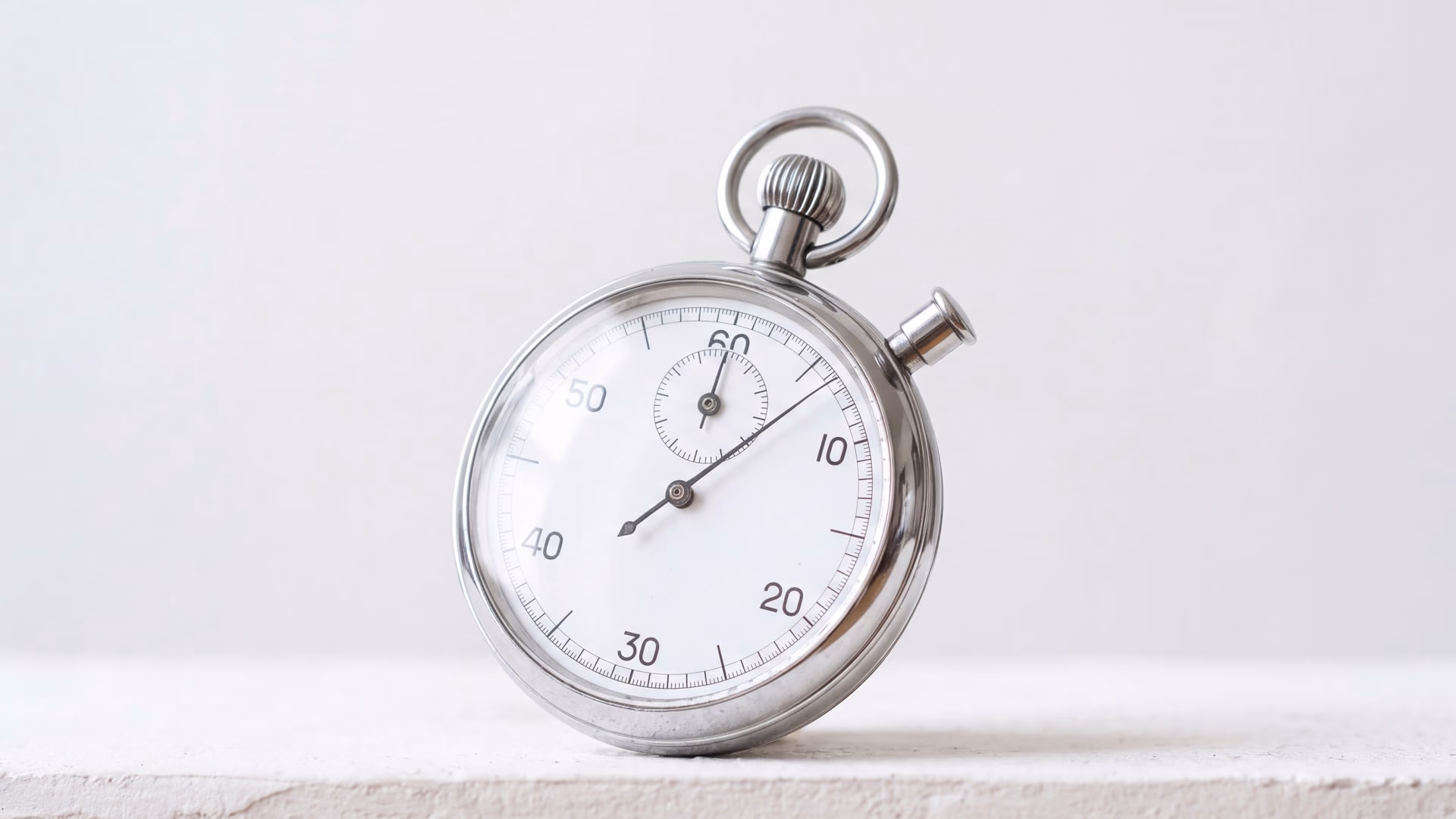 Un chronomètre mécanique posé sur un rebord de plâtre clair, cadran blanc et boîtier d'acier en pleine lumière du jour, aiguille des secondes en course.